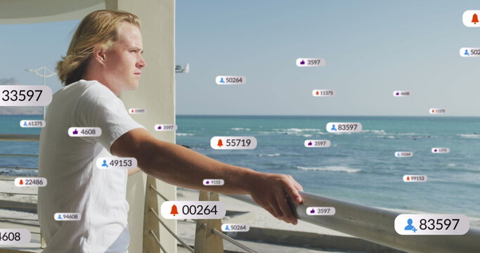 Multiple digital icons with increasing numbers over caucasian man enjoying the view from balcony