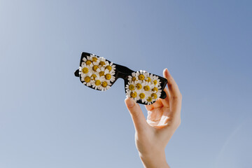 Hand holding sunglasses decorated with chamomile daisy flower buds over blue sky. Spring, summer sunny day concept with copy space for text - obrazy, fototapety, plakaty