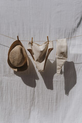 Female swimwear and straw hat hanging over white cotton cloth with strong shadows. Sunbathing on a summer sunny day concept - obrazy, fototapety, plakaty