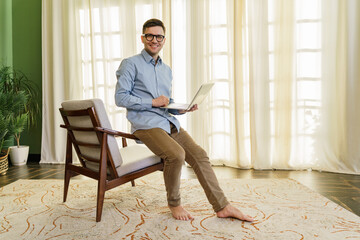 Cheerful barefoot man using a laptop in a sunny room with a green wall and sheer curtains.