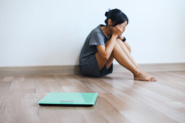 Weighing scale in front of sad young skinny woman sitting on floor due to unhealthy diet