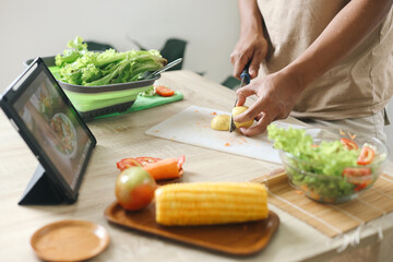 Man watching online culinary class on tablet, making salad with fresh vegetables