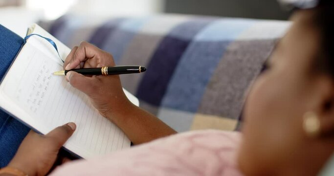 African American woman writes in a notebook, seated on a couch at home