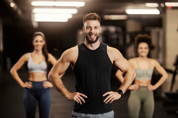 A strong sportsman is confidently posing in a gym and smiling at the camera. There are sportswomen in a blurry background.