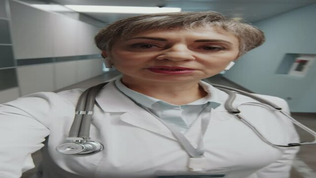 Vertical POV shot of mid-aged female doctor in white coat and wireless earphones going through hospital corridor and talking on camera during online video call