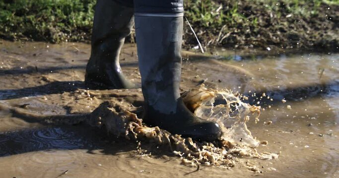 Super slow motion close up of farmer in rubber boots walks through mud puddles with splashing water drops after rainy day on green agricultural natural food production farmland fields at 1000 fps.