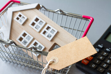 Miniature model of wooden house in shopping basket with tag and calculator, concept of selling, buying or renting real estate