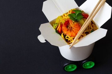 Chinese noodles with shrimps and vegetables in take-out box with car wheels on dark background. Creative chinese food delivery concept