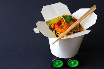 Chinese noodles with shrimps and vegetables in take-out box with car wheels on dark background. Creative chinese food delivery concept
