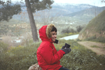 Smiling teenage traveler boy holding a thermos mug with hot drink, relaxing on a forest log while trekking in mountains