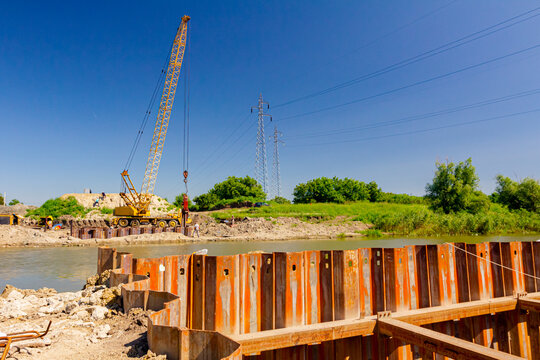 Bridge foundation made of metal piles on river coast in background is old hoist with pneumatic hammer. Building site area - Powered by Adobe