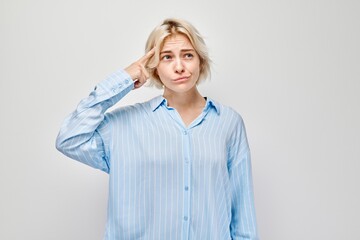 Confused young woman in blue shirt making finger gun gesture to head on a gray background.