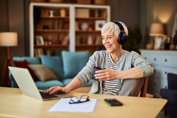 A happy senior adult woman working from home using laptop while enjoying a cup of coffee and...