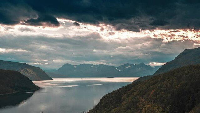 Rays of the setting sun pierce through the dense stormy clouds whirling above the calm fjord. A timelapse video.