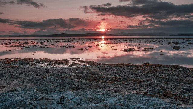 Dramatic sunset sky with red sun and dark clouds above the calm fjord at low tide in a timelapse video.