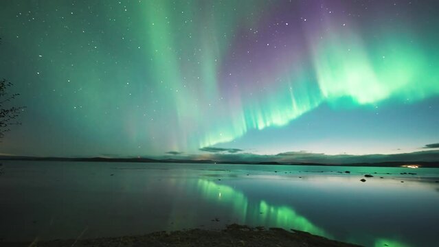 A magnificent light show of the Aurora Borealis in the night sky reflected in the calm waters of the fjord. A timelapse video.