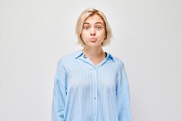 Confused young woman in blue shirt shrugging with puffed cheeks against a grey background.