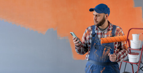 Painter man holding a paint roller and mobile phone sending a message on wall background, making...