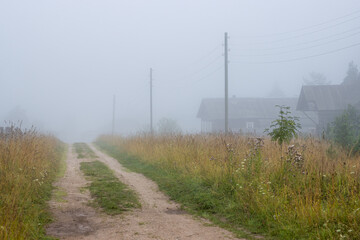 View of a deserted country dirt road and old wooden houses in the fog. Grass on the roadside. Misty morning in the countryside. Summer in the village. Foggy rural landscape. Vologda region, Russia.