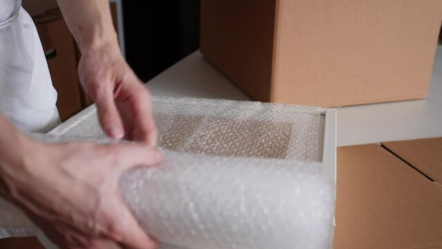 Close-up cropped shot of unrecognizable man packing photo with frame in bubble wrap ready to relocation to new home, cardboard box on table. Concept of new life in new home relocation, slow motion.
