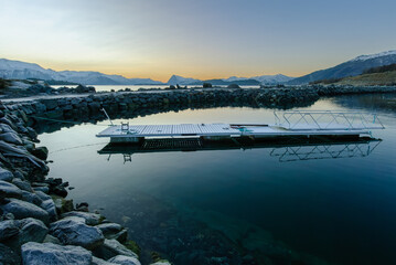 A tranquil scene as the sun sets behind snow-covered mountains, reflecting off the placid sea.