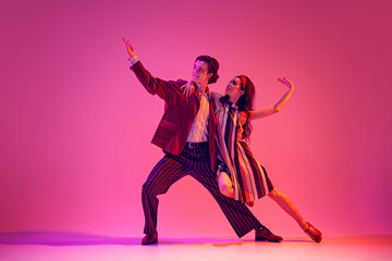 Artistic young man and elegant woman, couple dancing retro dance in stylish costumes against pink background in neon light. Concept of hobby, dance class, party, 50s, 60s culture, youth