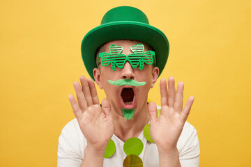 Shocked scared man wearing green hat and clover glasses standing isolated over yellow background...
