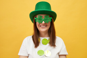 Cheerful delighted woman wearing green leprechaun hat and shamrock glasses posing isolated over...