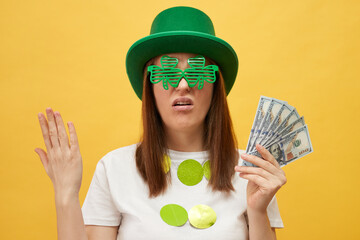 Displeased woman wearing green leprechaun hat and shamrock glasses posing isolated over yellow...