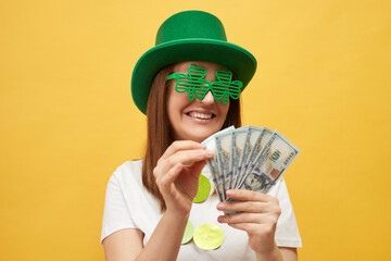 Smiling happy woman wearing green hat and clover glasses standing isolated over yellow background...
