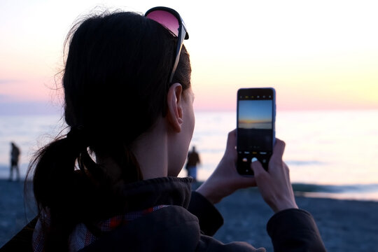 Woman tourist taking photoon phone of sea sunset admiring scenery nature, back view. Sundusk, dark evening on seashore. Female enjoying pastime on coast on vacation. Travel, tourism, journey concept.