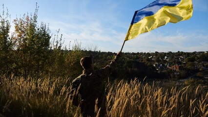 Male military in uniform waving flag of Ukraine at countryside. Young soldier of ukrainian army lifting blue-yellow banner as symbol of victory against russian aggression. Invasion resistance concept. - 757185270