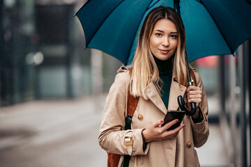 Fashionable smiling young woman under an umbrella holding a mobile phone.