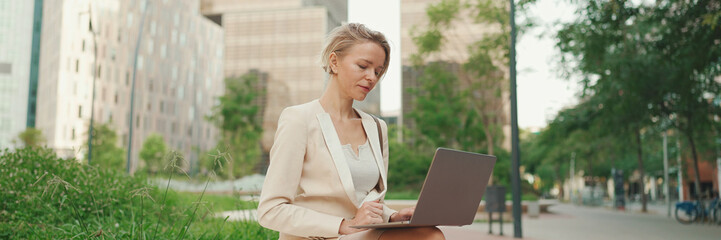 Smiling businesswoman with blond hair wearing beige suit is using pc laptop outside, Panorama