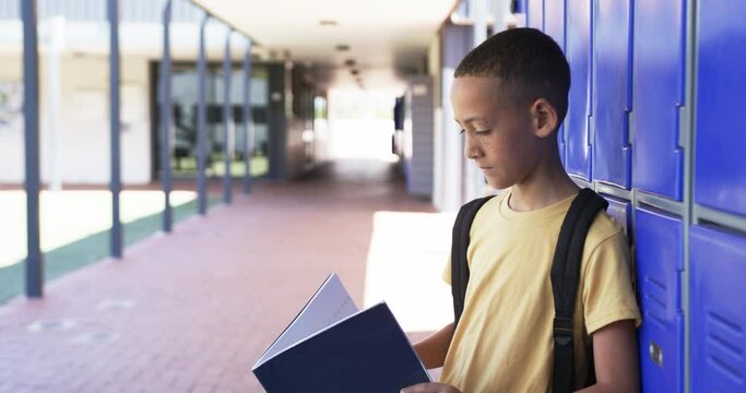 Biracial boy with a backpack stands by blue lockers in school, reading a book, with copy space