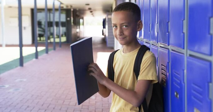 Biracial boy with a backpack stands by blue lockers, holding a folder, in school