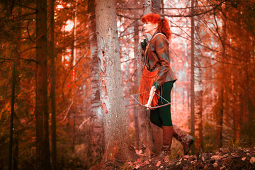 Mature model portraying a royal huntress with red curve hair is hunting with a crossbow in the in vibrant autumn forest in a thematic photo shoot - obrazy, fototapety, plakaty