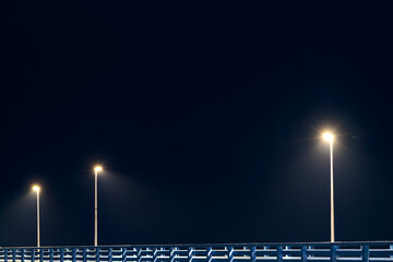 Street lampposts casts dim cold blue glow along shore pier standing against pitch black canvas of night sky paints poetic picture of pier in stillness of night, serenity of solitary moments - obrazy, fototapety, plakaty