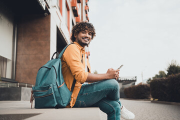 Photo of positive glad man sitting outdoors dormitory or university building spending free time reading scrolling instagram