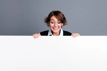 Deurstickers Photo of excited cheerful glad woman dressed stylish clothes peeking out white poster empty space isolated on grey color background © deagreez
