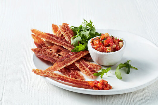 Crispy bacon strips served with a side of fresh salsa and garnished with arugula on a white plate.