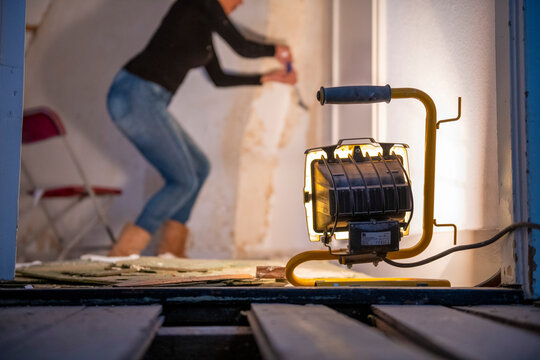 A focused individual works on a wall renovation illuminated by a bright portable work light.