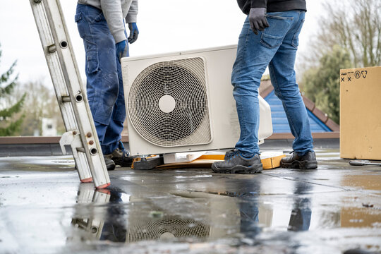Two workers installing an air conditioning unit on a rooftop on a cloudy day.
