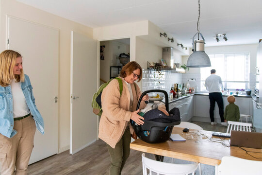 A family moment in a cozy kitchen with a woman showing a baby in a car seat to a smiling visitor