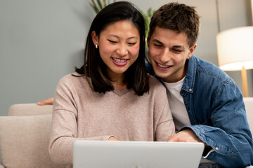 Happy young relaxed diverse couple sitting on sofa using laptop. Affectionate excited woman and man using computer at living room.