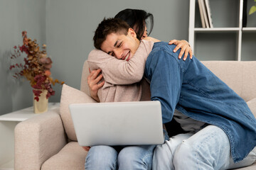 Young excited diverse couple hugging while working with laptop. Female and male romantic partners working online and celebrating successful on a sofa.