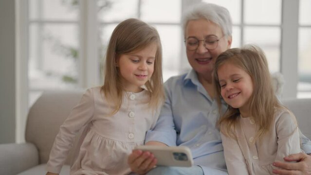 Grandmother with granddaughters resting. Attractive relaxed older adult granny embracing watches cartoons on smartphone with children two twins leisure rest at home. Family love, generation concept.