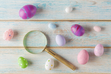 Top view of a magnifying glass  next to colorful, textured eggs on a rustic wooden surface. Easter eggs hunting and search - 757242883