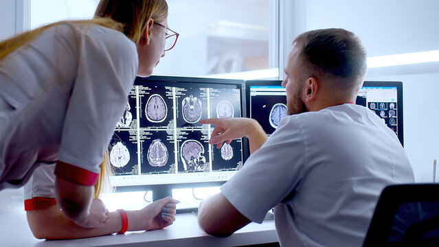 In Control Room Doctor and Radiologist Discuss Diagnosis while Watching Procedure and Monitors Showing Brain Scans Results, In the Background Patient Undergoes MRI or CT Scan Procedure.