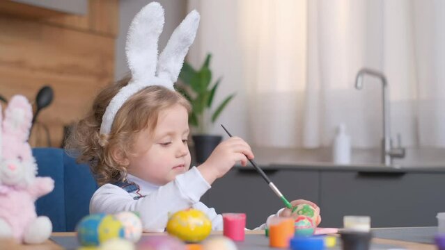 A toddler with bunny ears is happily painting Easter eggs at a table, sharing a smile while enjoying leisure time 